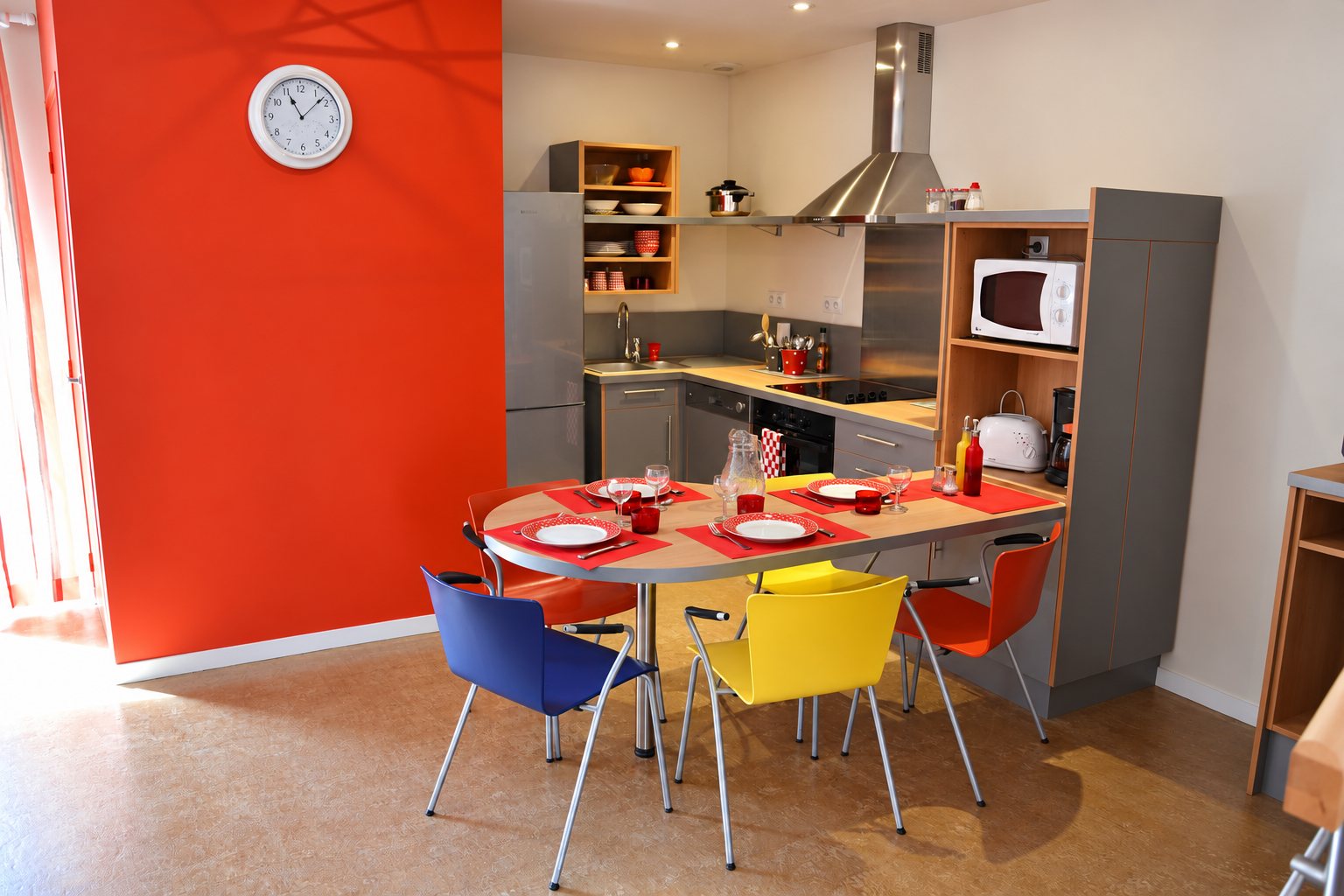 Une cuisine moderne aux murs orange, avec une table ovale dressée pour quatre et des chaises bleue, jaune et orange.