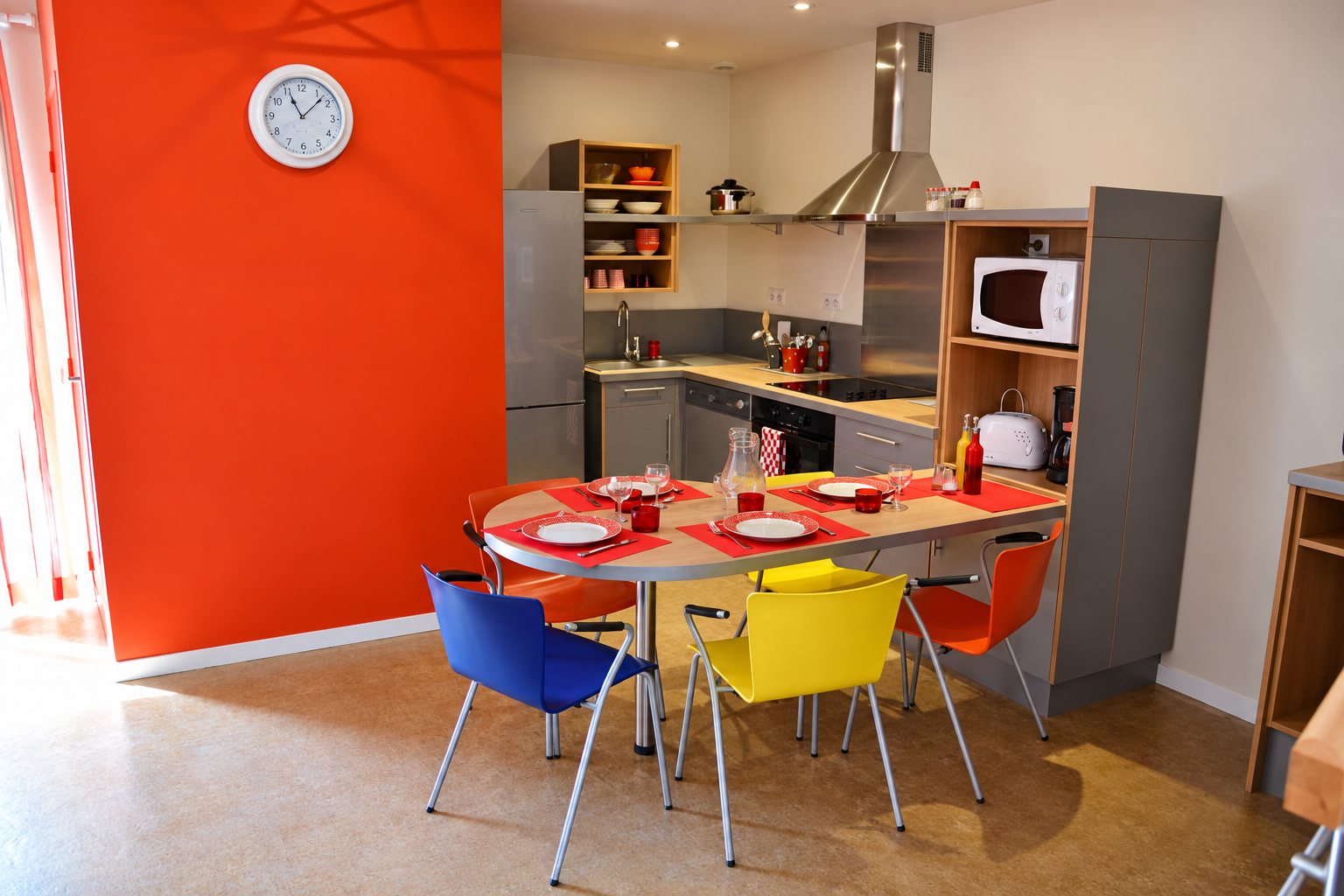 Une cuisine moderne avec un mur orange, une table ovale dressée pour quatre et des chaises bleue, jaune et orange.