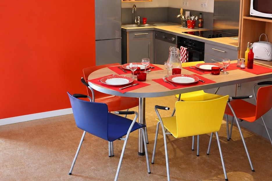 La table ovale dressée pour quatre, avec ses chaises bleue, jaune et orange, devant le mur orange.