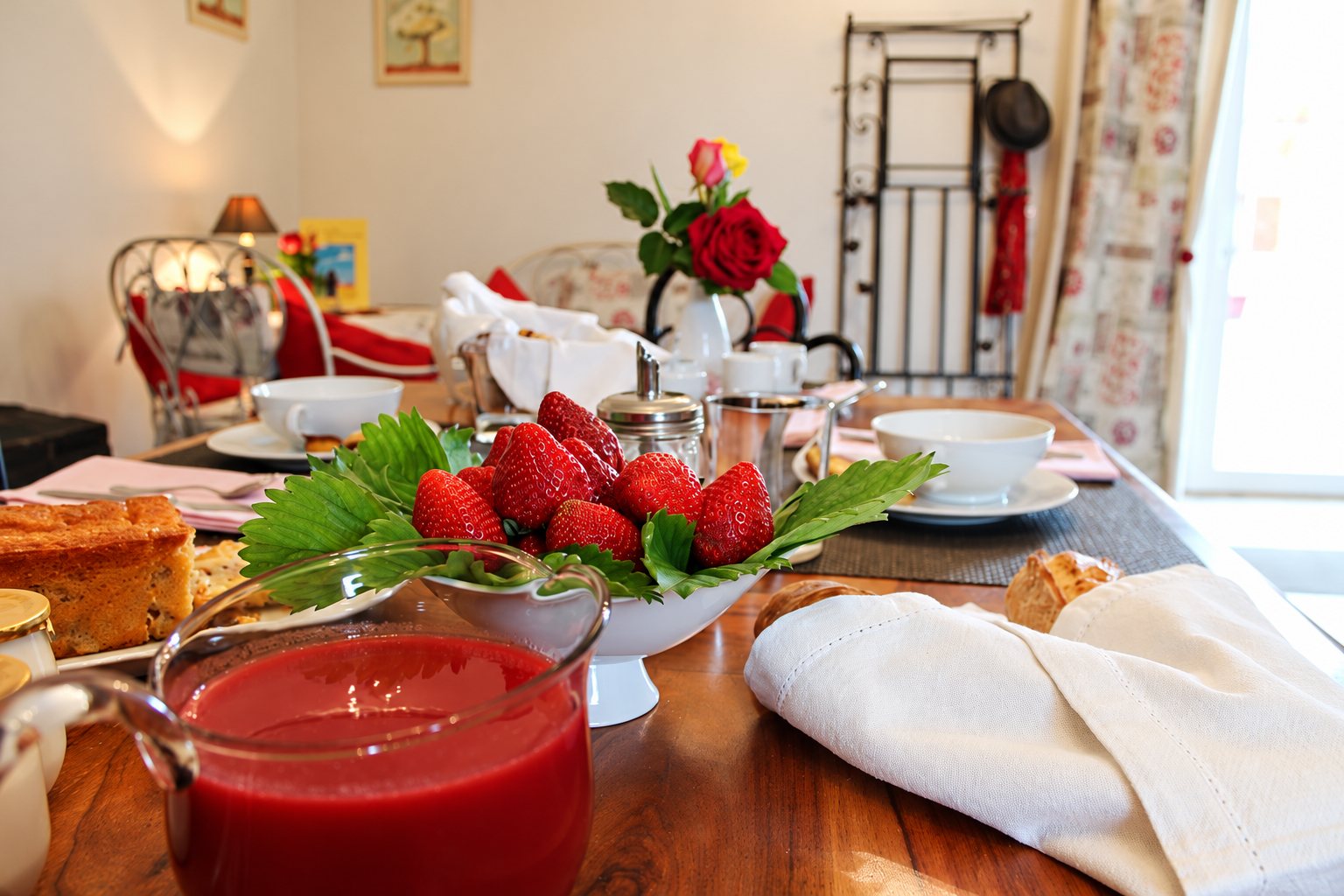 Une table de petit-déjeuner dressée avec un bol de fraises fraîches, un pichet de coulis rouge, du gâteau, du pain et un bouquet de roses en arrière-plan.